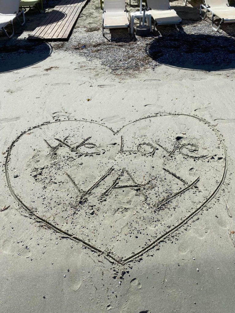 Egy szív alakzatot rajzolnak a homokba egy tengerparton, benne a "We love VAY" felirattal. A strandon elszórtan hínár és kagyló található. A háttérben nyugágyak és fa emelvények láthatók.
