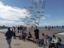Az emberek összegyűlnek és leülnek az "Esernyők" szobor közelében, amely George Zongolopoulos szabadtéri művészeti installációja a kék ég és a tenger hátterében. Vannak, akik sétálnak, míg mások beszélgetnek és pihennek a környéken.