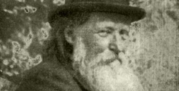 Egy régi szürkeárnyalatos fénykép egy idős férfiról, hosszú fehér szakállal, sötét, karimájú kalapot és kabátot visel. Arckifejezése nyugodt, ahogy kissé oldalra néz az elmosódott, rusztikus háttér előtt.