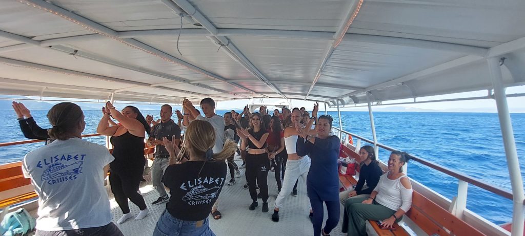 Egy nyüzsgő embercsoport táncol egy hajón a fedett fedélzet alatt egy napsütéses napon. A hajó kint van a tengeren, kék vizekkel és tiszta égbolttal a háttérben. Az előtérben többen "Elisabet Cruises" feliratú inget viselnek.