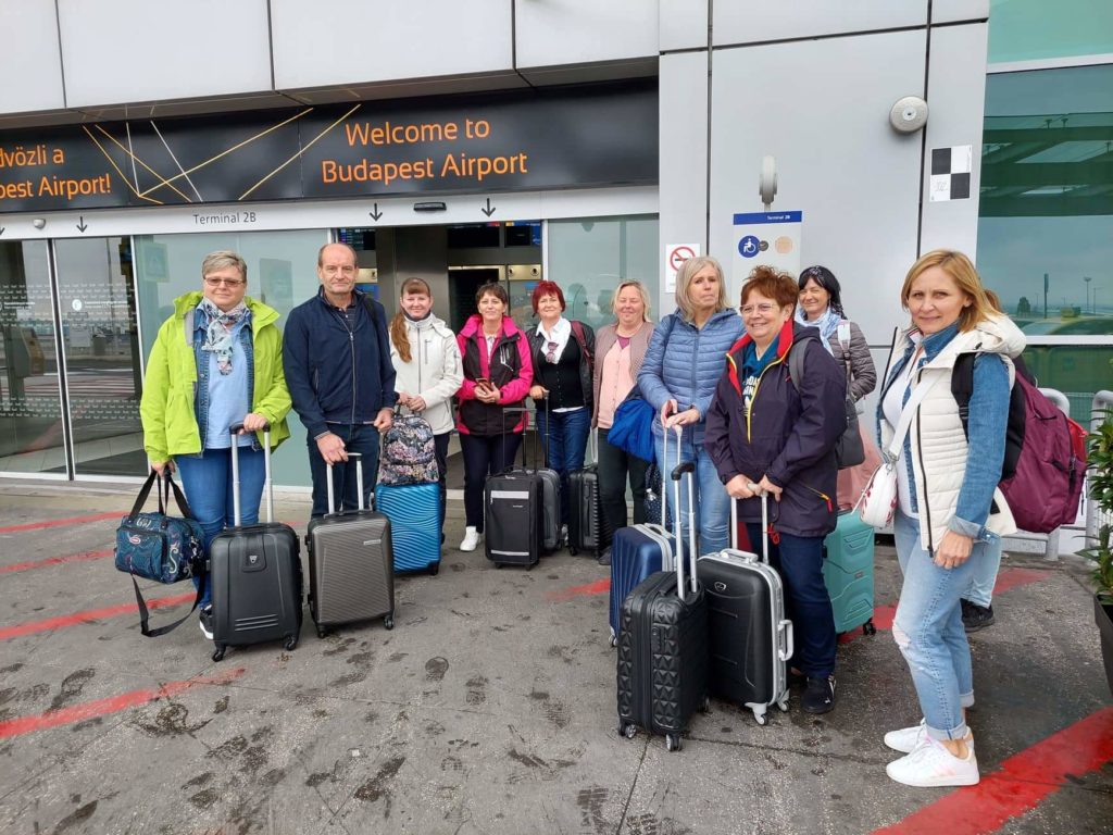 Tizenkét fős csoport poggyászokkal áll a Budapest Airport bejárata előtt. Alkalmi utazóruhába vannak öltözve, zakókkal és kabátokkal. Az ég borúsnak tűnik, és a 2B terminál tábla közelében gyűltek össze.