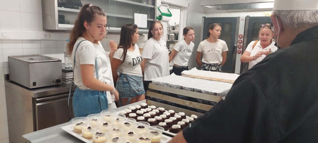 Egy csapat fiatal nő áll a konyhában, fehér felsőt és kék farmert visel. Egy fekete inges, fehér sapkás szakácsot hallgatnak, aki hozzájuk beszél. A konyhapulton tálcák találhatók a desszertekhez, beleértve a süteményt és a pudingos csészéket.