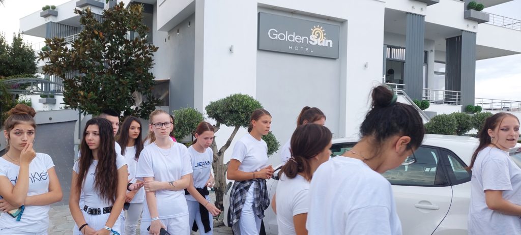 A Golden Sun Hotel előtt fehér ingbe öltözött fiatalokból álló csoport áll, főként lányok. Egyesek papírokat tartanak a kezében, mások pedig csevegnek vagy a telefonjukat nézik. A háttérben fák és a szálloda épületének modern homlokzata látható.