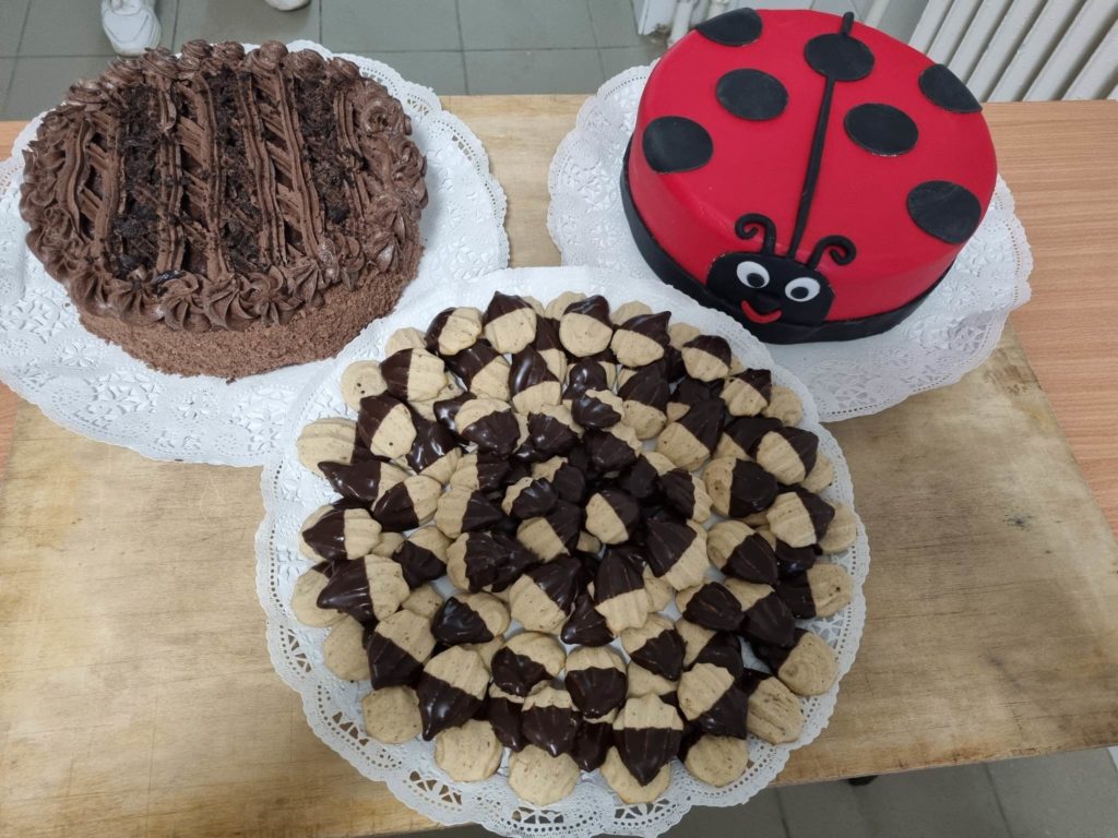 Három desszert látható egy fa felületen. Az első egy csokitorta krémes díszítéssel. A második egy katicabogár témájú torta piros és fekete fondanttal. A harmadik egy tálca süti, félig csokoládéval bevonva és szépen elrendezve.