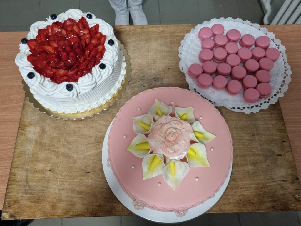 Fafelületen három torta látható: egy eper torta tejszínhabbal és áfonyával, egy rózsaszín torta virágdíszítéssel a közepén és egy tál rózsaszín macaron.