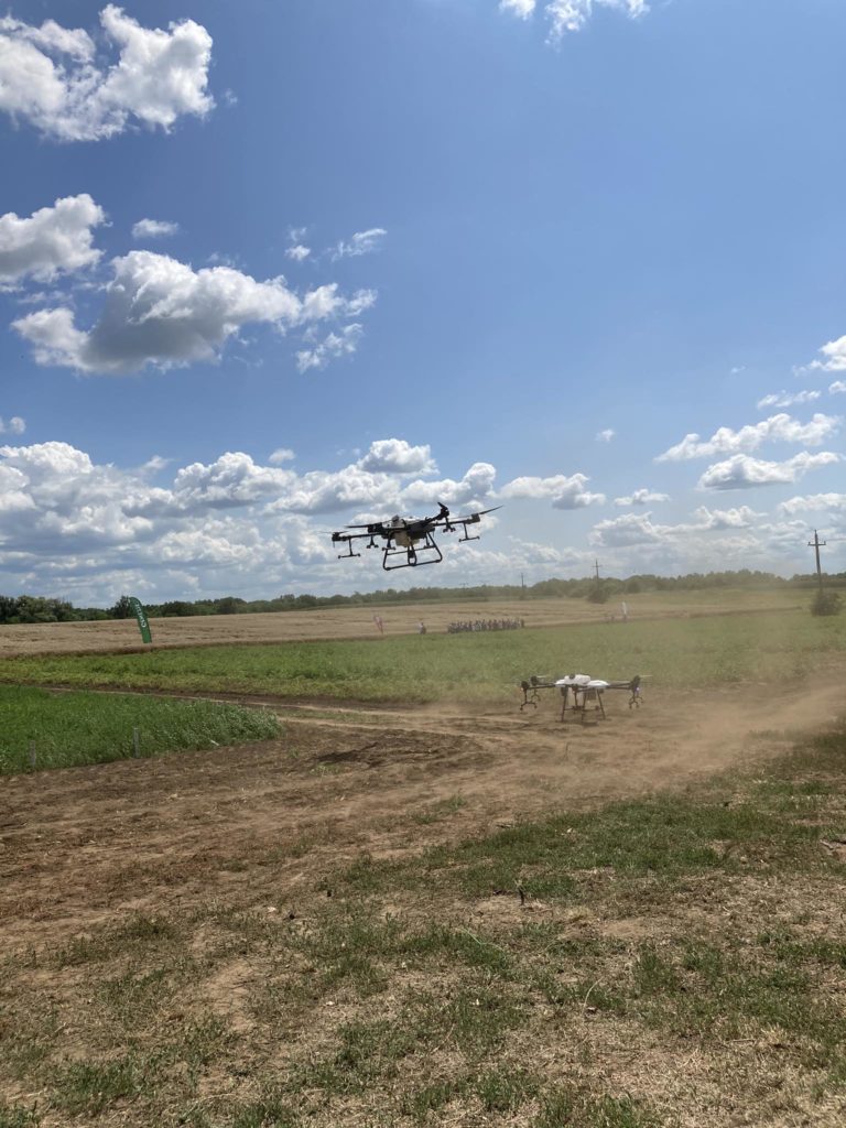 Egy drón repül egy poros mező felett, néhány emberrel a távolban. Az ég többnyire derült, néhány bolyhos felhővel, a környék pedig zöldellő füves foltokkal. A jelenet világos és napos, ami kellemes napot sugall.