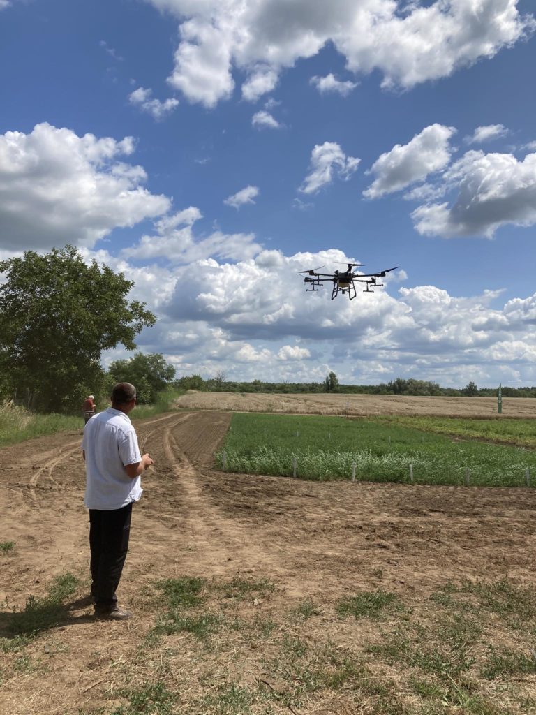 Egy fehér inges ember drónt üzemeltet egy vidéki mezőn egy napsütéses napon, kék éggel és szétszórt felhőkkel. Egy másik személy hátrébb áll és figyeli. A területet zöldövezet és földút veszi körül.