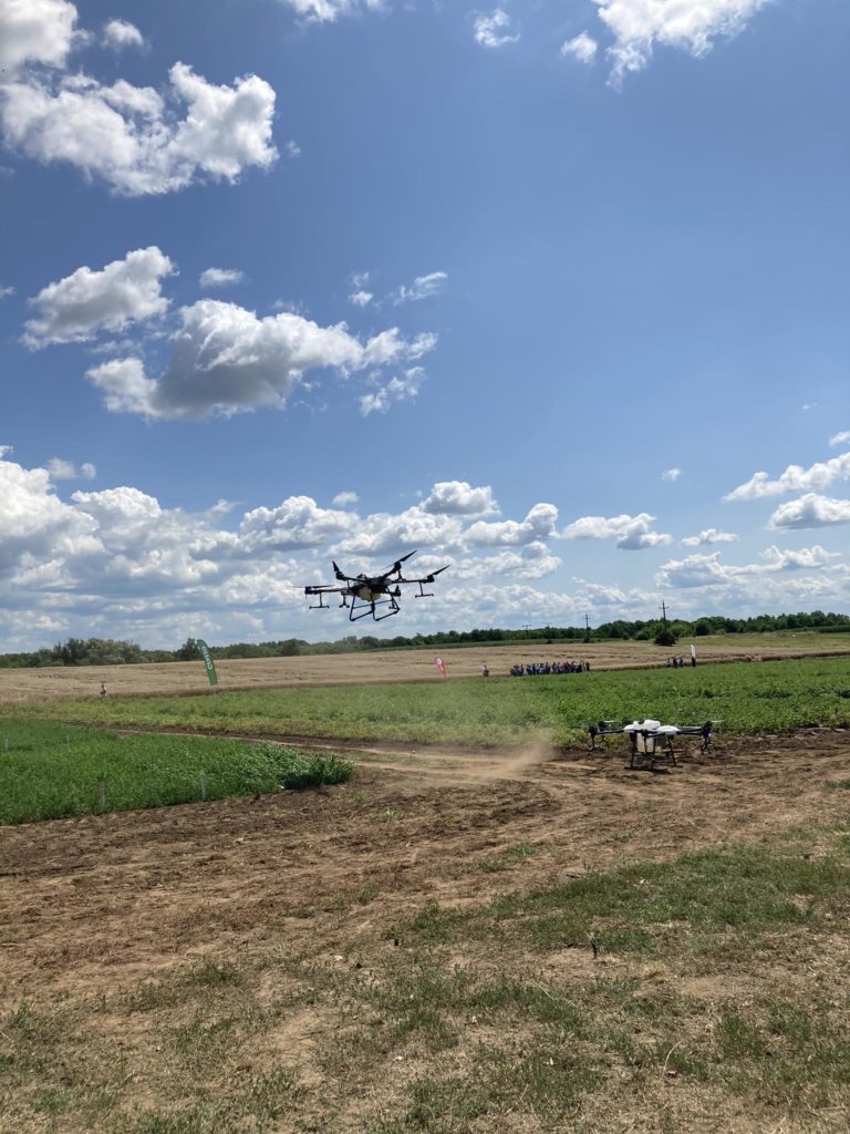 Egy nagy drón repül egy zöld és barna mezőgazdasági mező felett a kék ég alatt, szétszórt felhőkkel. Egy kis embercsoport áll a háttérben, közel a mező széléhez.