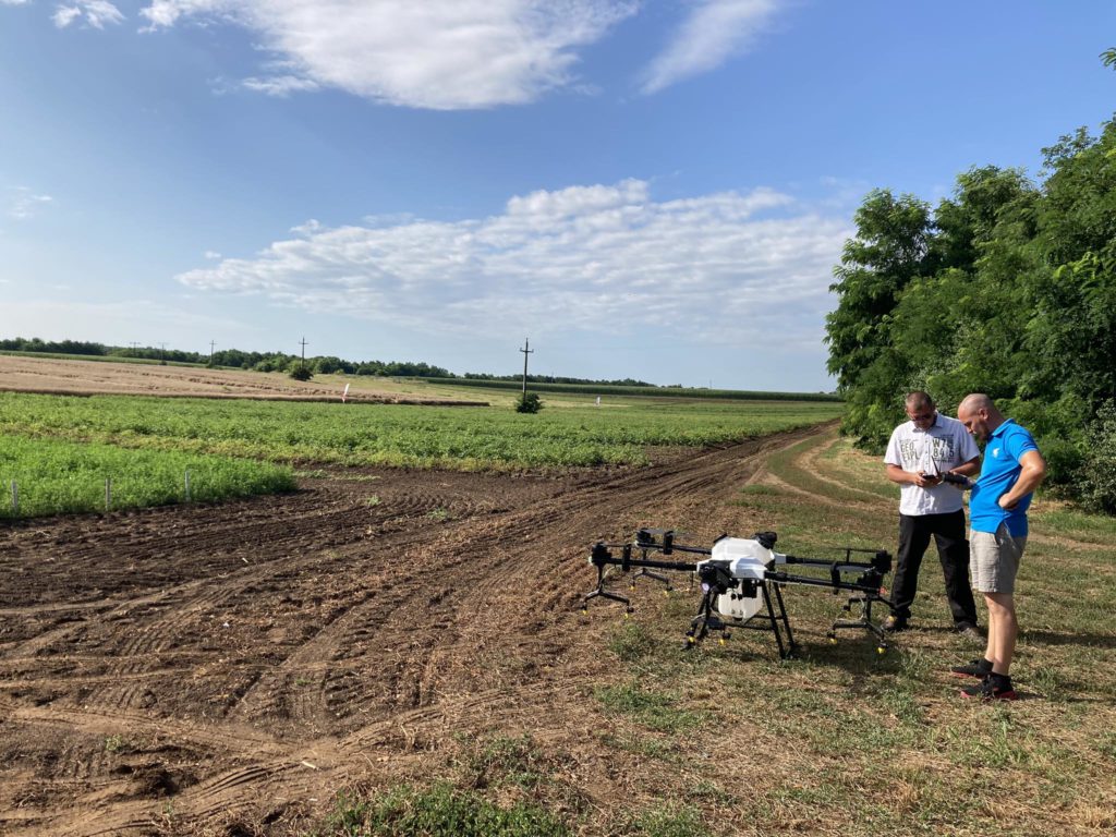Két személy egy vidéki földúton áll, és egy táblagépet vizsgál. Előttük egy nagy mezőgazdasági drón. A háttérben kiterjedt, megművelt területek láthatók a részben felhős égbolt alatt, napfényfoltokkal. Jobb oldalon fák láthatók.
