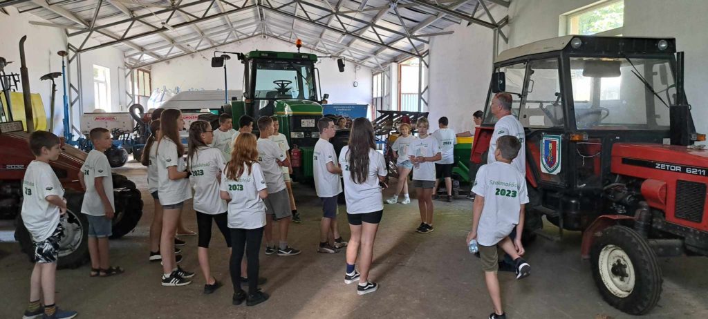 Egy nagy garázsban vagy műhelyben összegyűlt egy csapat gyerek, akik hozzáillő fehér pólót viseltek, hátulján „Słodkiewicz 2023” felirattal. Különféle traktorok és mezőgazdasági gépek veszik körül őket, egy férfi beszél hozzájuk egy piros traktor előtt.