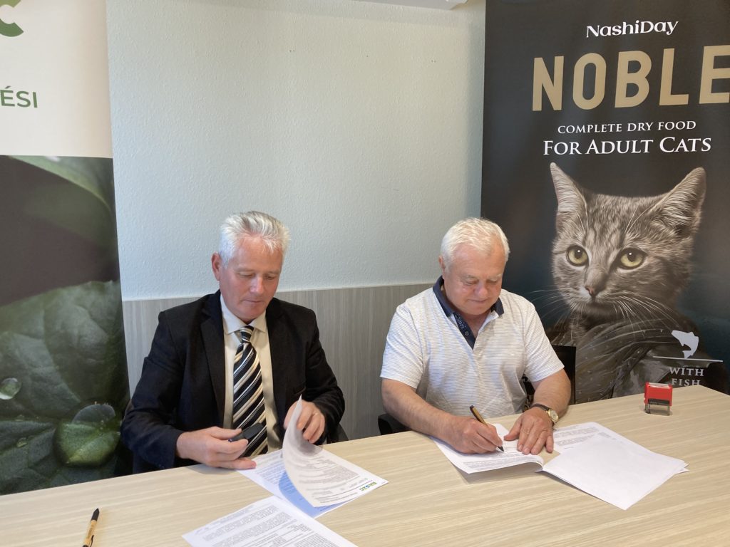 Két férfi ül egy asztalnál és dokumentumokat ír alá. Az egyik férfi fekete öltönyt és csíkos nyakkendőt, míg a másik fehér, rövid ujjú inget visel. Mögöttük transzparensek vannak, az egyiken egy macska képe és a „Nemes teljes értékű szárazeledel felnőtt macskáknak” szöveggel.