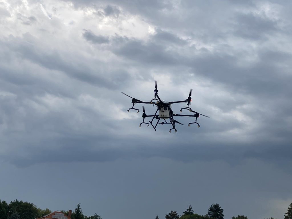 Egy nagy drón több rotorral repül az égen egy felhős napon. A háttérben a fák teteje és épületrészek láthatók.