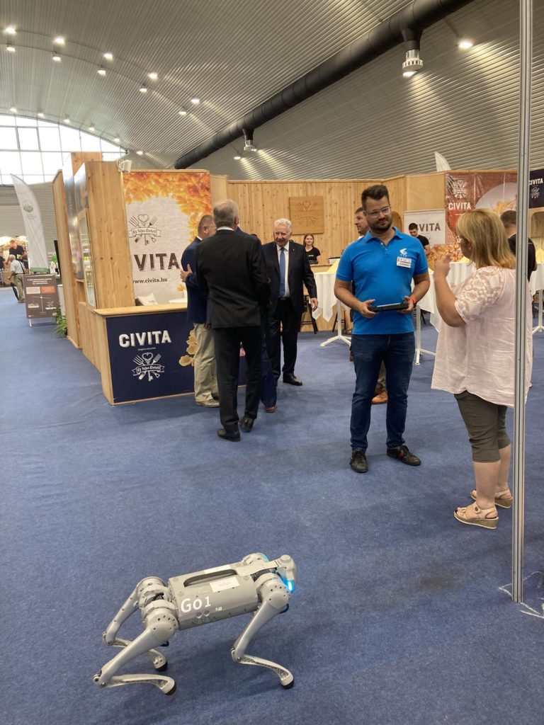 Egy csoport ember, köztük egy idősebb férfi öltönyben, két férfi üzleti öltözékben és egy nő hétköznapi ruhában, egy kiállítási standban áll. Az előtérben egy kis négylábú robot látható, „G01” felirattal. A standon a "Civita" márkajelzés található.