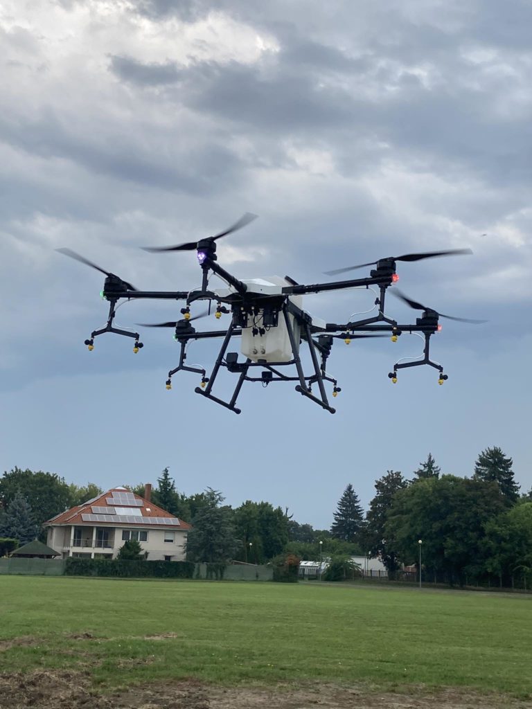 Egy nagy drón több rotorral lebeg a levegőben egy nyílt mező felett. A háttérben fák, napelemes ház a tetőn és felhős égbolt látható. A drón különféle tartozékokkal és lámpákkal rendelkezik, amelyek jelzik működési állapotát.