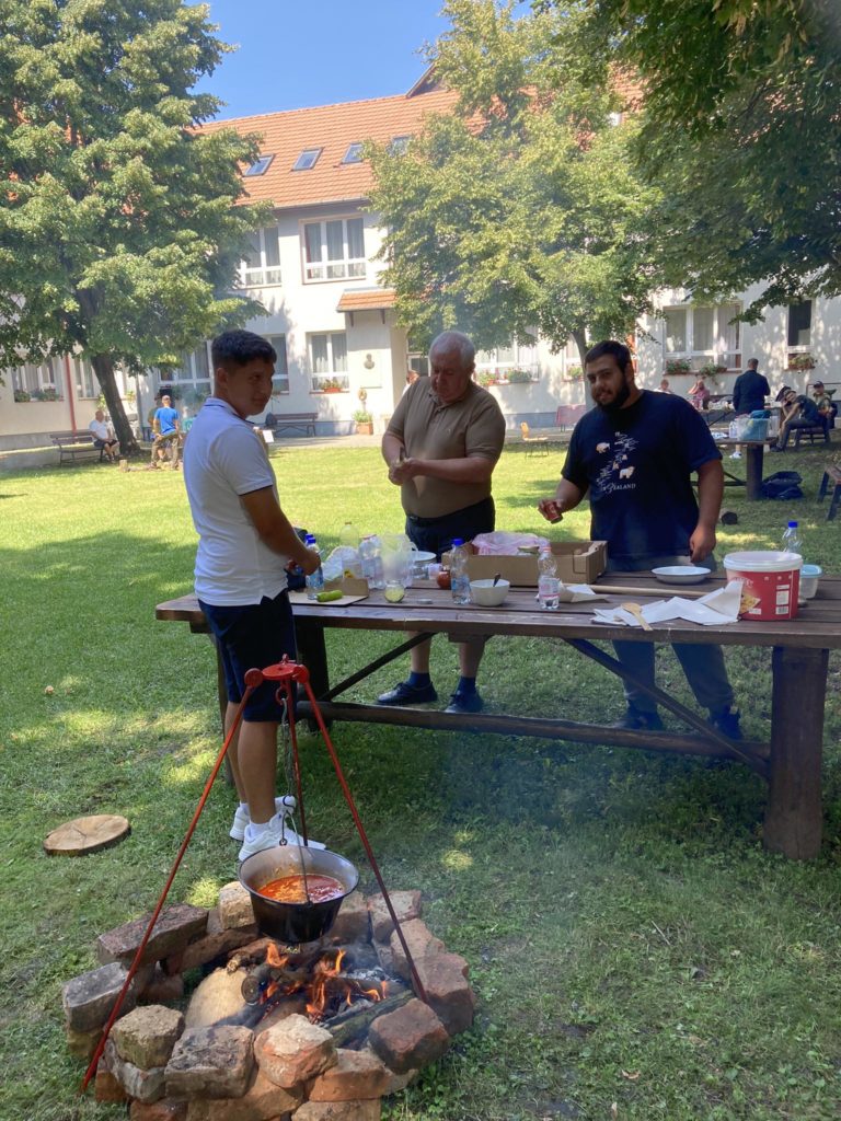 Három férfi a szabadban főz egy piknikasztalnál a pázsiton. Különféle főzőeszközök és edények vannak az asztalon. Egy edény lóg előttük egy kis tábortűz fölött. A háttérben többen ülnek és sétálnak. Fák és épületek láthatók.
