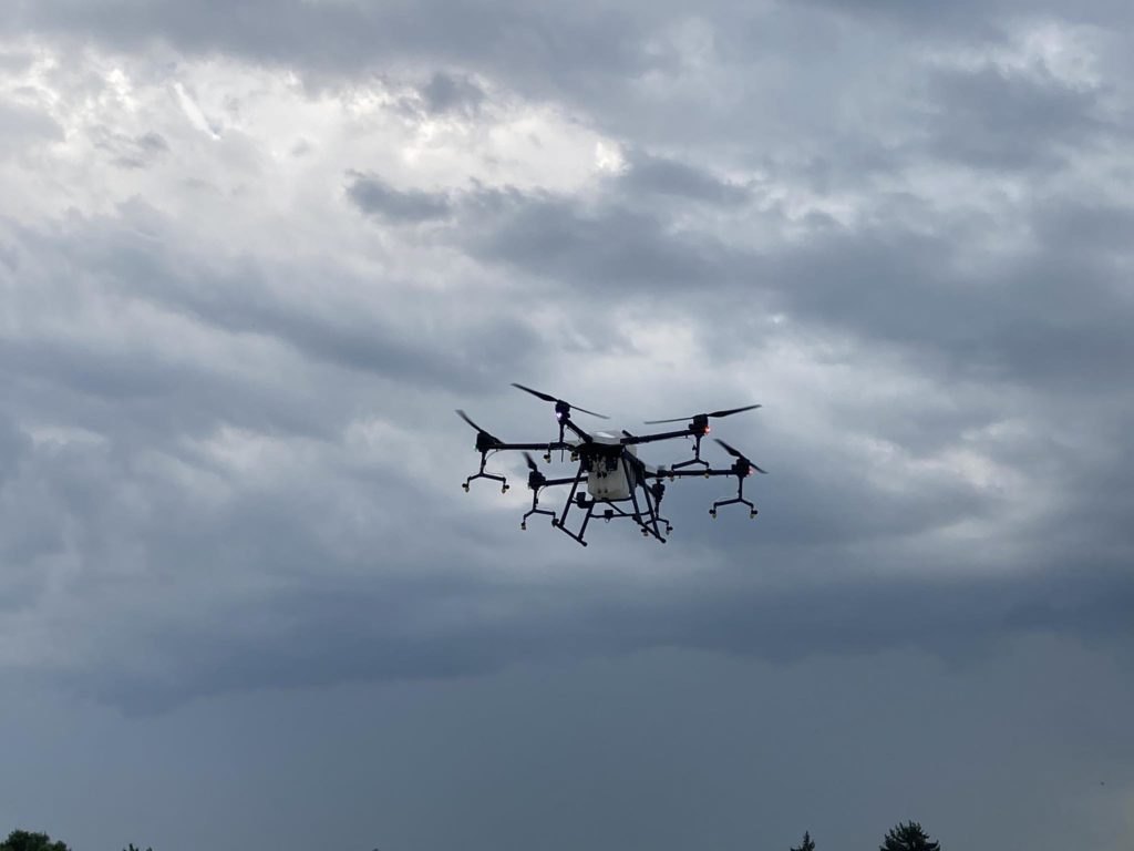 Egy drón lebeg az égen a szürke, felhős időjárás hátterében. A kép alján halványan látszik a lenti táj, néhány fa teteje kikandikál. A drónnak több légcsavarja és központosított teste van.