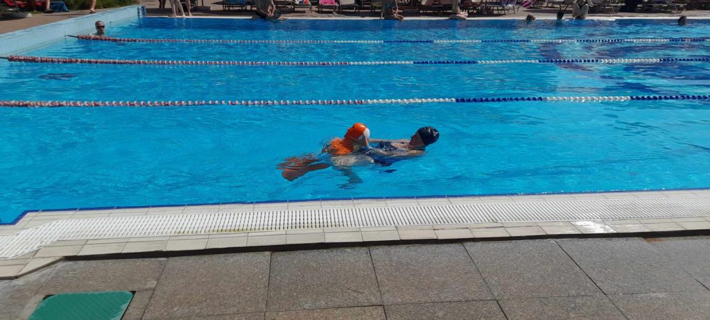 Egy kék úszósapkát és narancssárga karját viselő személy úszik egy tiszta kék úszómedence egyik sávjában. A medencének több sávja van sávkötelekkel, és a háttérben más emberek úsznak és heverésznek. A medencepart járólapos kőből készült.