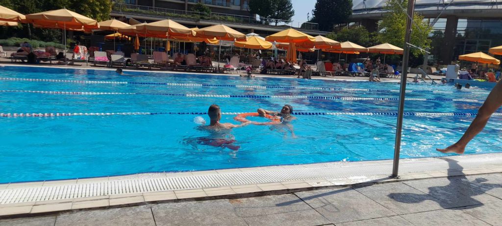 Három ember úszik egy ragyogó kék vizű szabadtéri medencében. A medence szélén narancssárga napernyők és nyugágyak sorakoznak, alattuk az emberek pihennek. A háttérben fák és épületek láthatók, kellemes hangulatot teremtve.