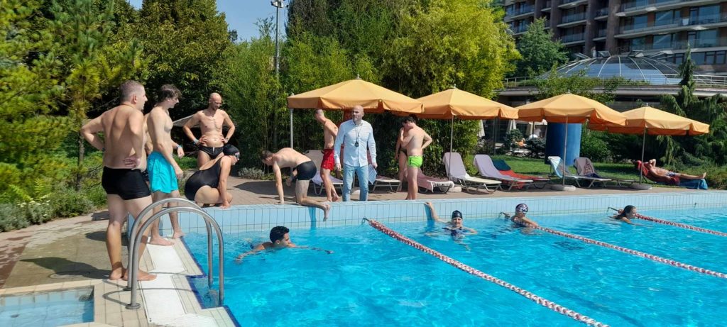 Emberek csoportja gyűlt össze egy szabadtéri medence körül egy napsütéses napon. Vannak, akik úsznak, míg mások a medence mellett állnak vagy ülnek, és fürdőruhát viselnek. A háttérben sárga esernyős nyugágyak és zöld fák állnak.