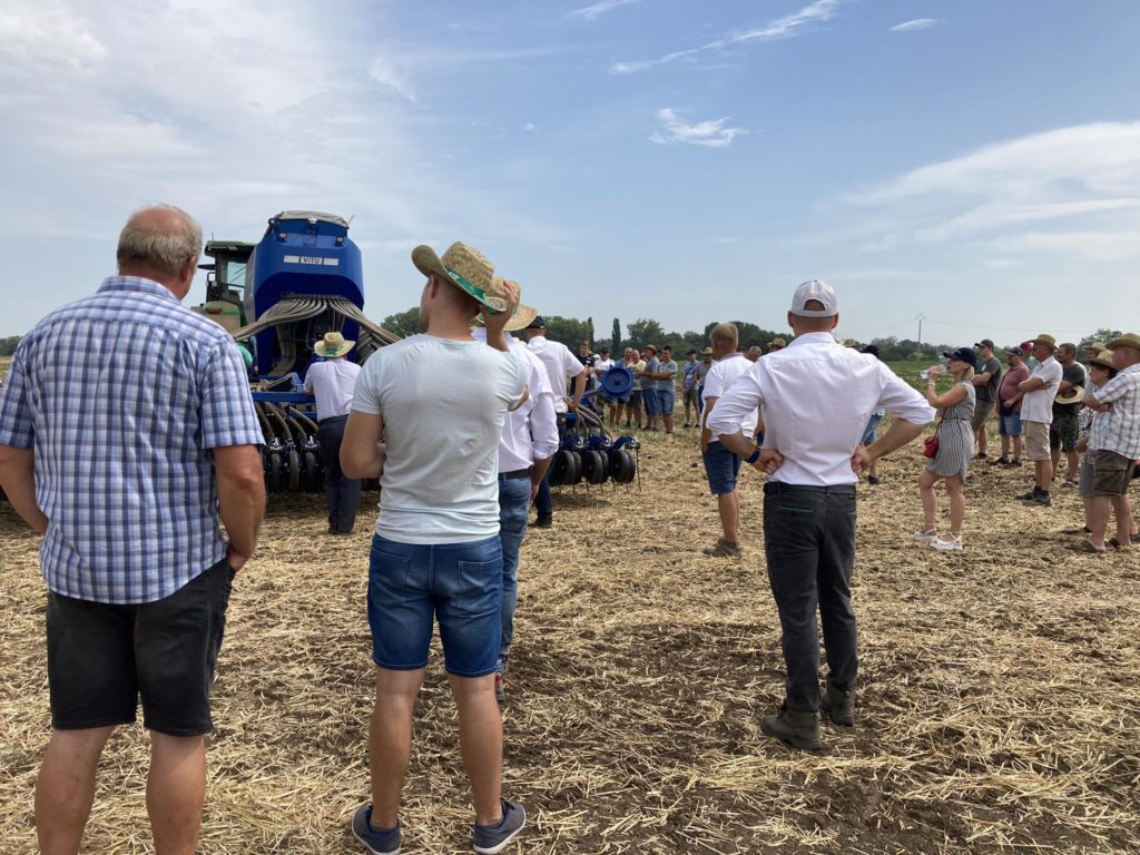 Egy csoport ember áll a mezőn, és figyelmesen néz egy kék mezőgazdasági gép bemutatót. Vannak, akik kalapot és alkalmi nyári ruhát viselnek. A mezőn száraz a szalma, és részben felhős az ég.