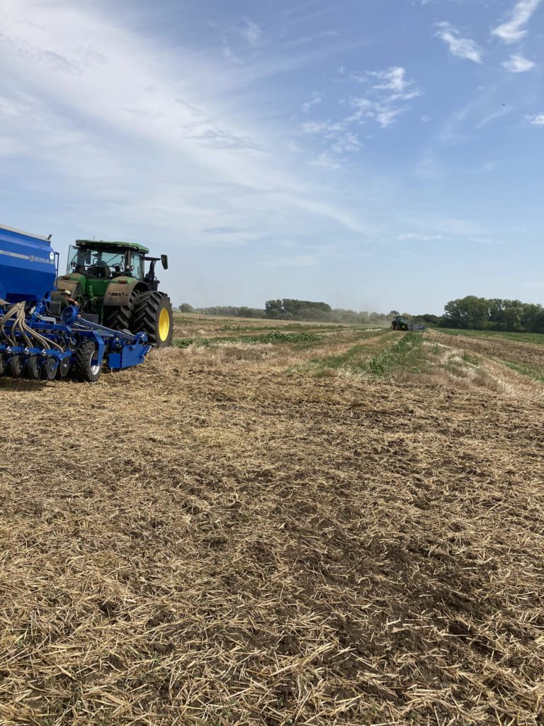 Egy zöld traktor egy kék mezőgazdasági gépet húz egy hatalmas, frissen művelt szántóföldön, részben felhős égbolt alatt. A távolban egy másik traktor üzemel, zölddel és fákkal körülvéve. A jelenet mezőgazdasági tevékenységet és mezőgazdasági munkát ábrázol.