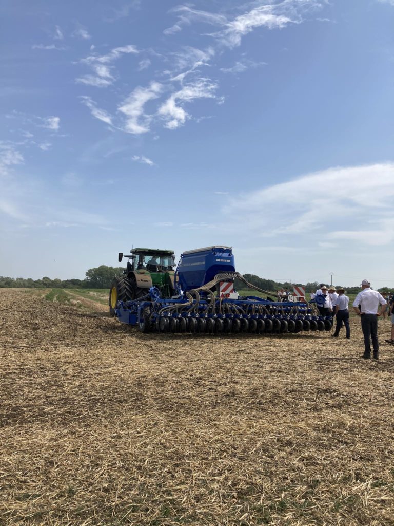 Egy nagy kék mezőgazdasági gép egy traktorhoz van rögzítve a nyílt területen. Többen fehér ingben és sötét nadrágban állnak a közelben, és figyelik a felszerelést. Tiszta az ég, szórványosan felhők.