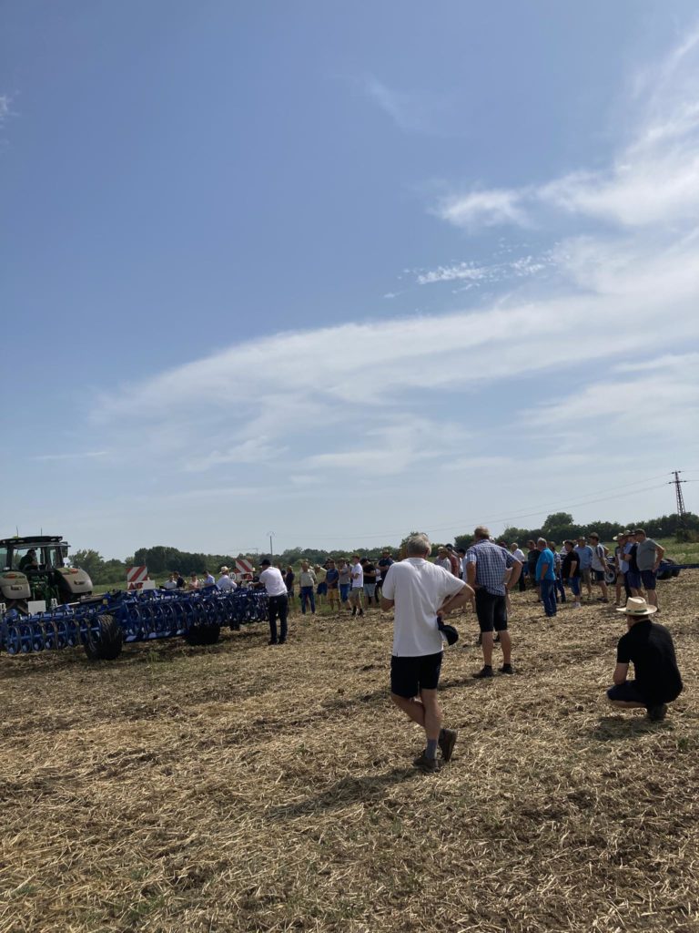 Egy csoport ember összegyűlik a szabadban egy mezőn, tiszta ég alatt. A bal oldalon láthatók a mezőgazdasági berendezések, beleértve a traktort és a kék mezőgazdasági gépeket. Néhány résztvevő áll, míg mások ülnek vagy térdelnek a földön, és figyelik a jelenetet.