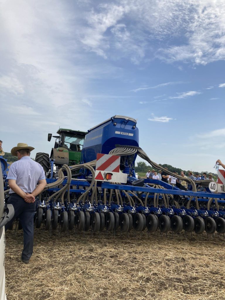 Az emberek nagy mezőgazdasági gépek, köztük egy kék és zöld traktor körül gyűlnek össze egy mezőn egy napsütéses napon. A berendezésben több sor fémtárcsa és tömlő található. Az előtérben egy kalapos és csíkos inges személy látható.