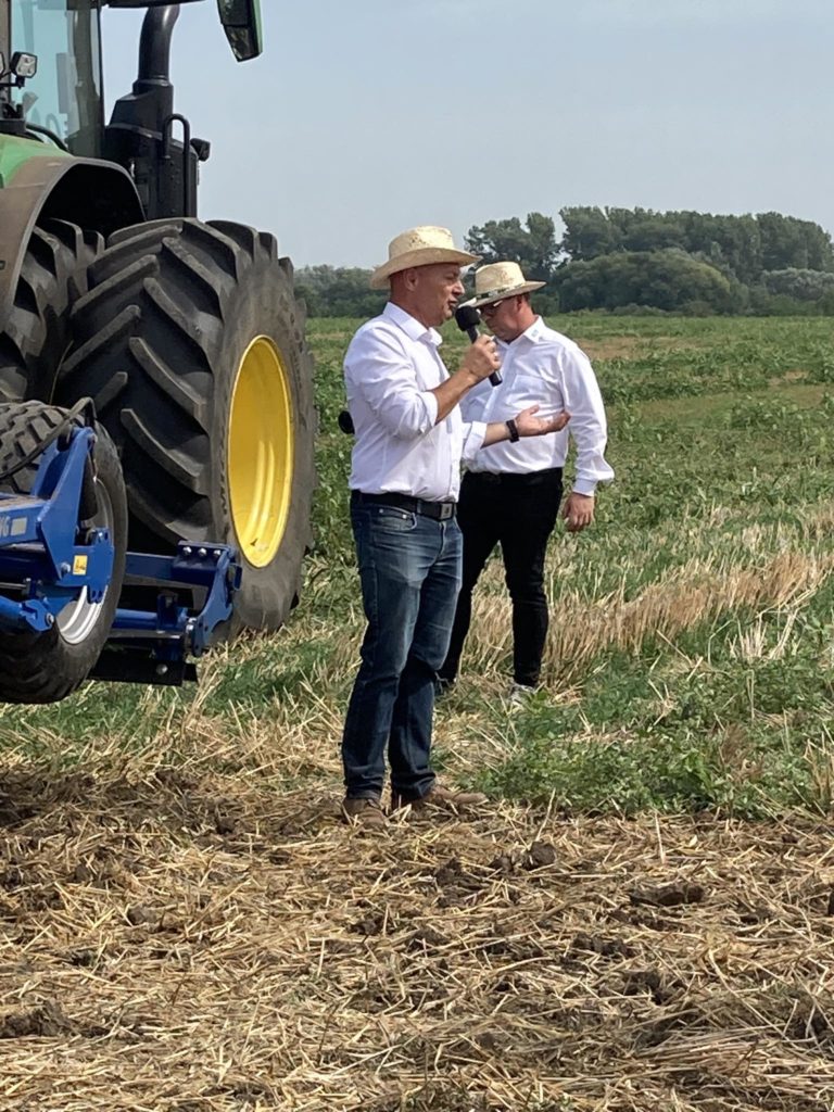 Két fehér inges és cowboykalapos férfi áll a mezőn egy nagy zöld traktor mellett. Az egyik férfi a mikrofonba beszél, míg a másik mögötte áll. Úgy tűnik, hogy egy összejövetelhez szólnak, vagy bemutatót tartanak mezőgazdasági környezetben.