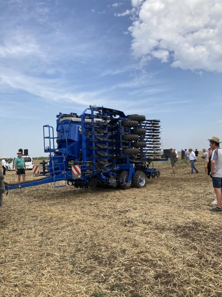 Tiszta időben az emberek egy nagy, kék mezőgazdasági gép körül állnak a mezőn. Úgy tűnik, hogy a gép egy nehéz mezőgazdasági berendezés, több kerékkel és talaj-előkészítéshez vagy ültetéshez szükséges gépekkel. Vannak, akik kalapot és alkalmi nyári ruhát viselnek.