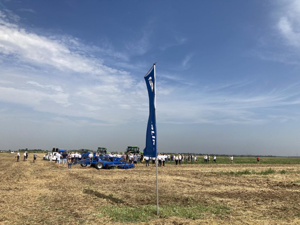 Emberek csoportja gyűlik össze egy nagy nyílt mezőn a kék ég alatt, szétszórt felhőkkel. Számos mezőgazdasági gép és traktor található. Az előtérben egy kék függőleges zászló, fehér szöveggel uralja a jelenetet. Úgy tűnik, az esemény a szabadban zajlik.