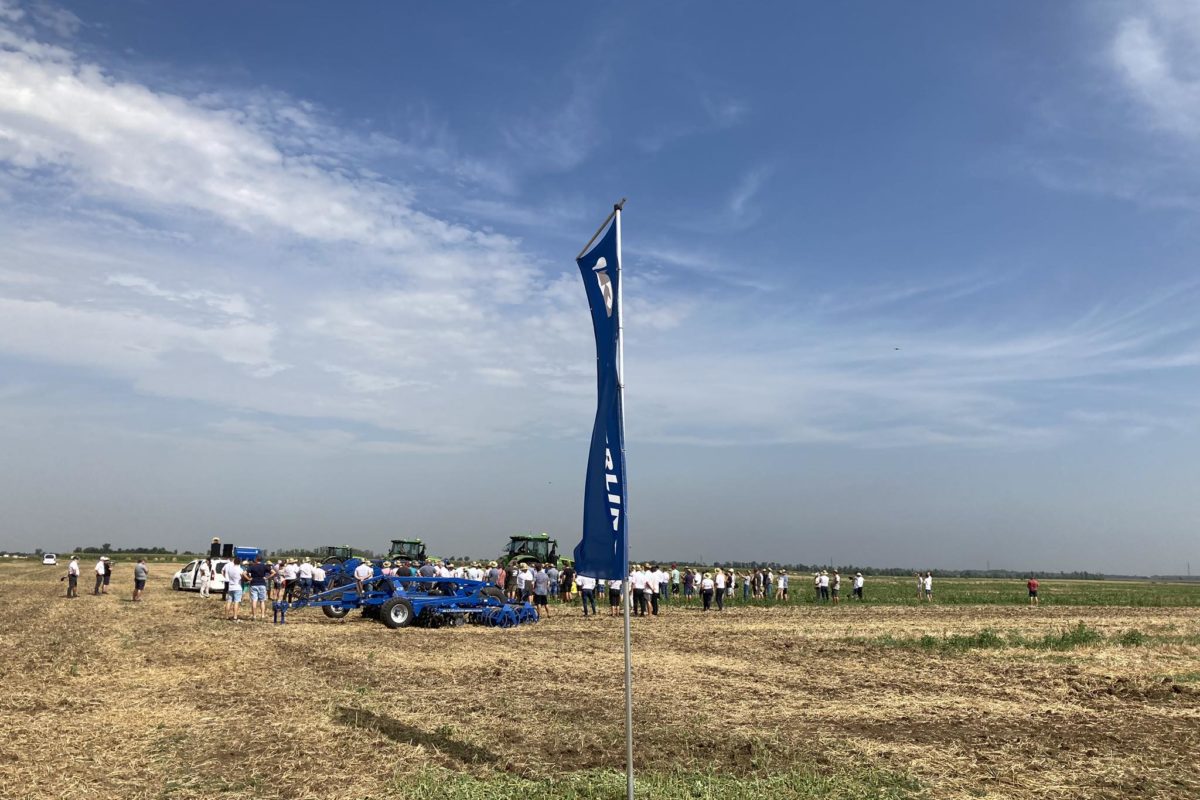 Emberek csoportja gyűlik össze egy nagy nyílt mezőn a kék ég alatt, szétszórt felhőkkel. Számos mezőgazdasági gép és traktor található. Az előtérben egy kék függőleges zászló, fehér szöveggel uralja a jelenetet. Úgy tűnik, az esemény a szabadban zajlik.