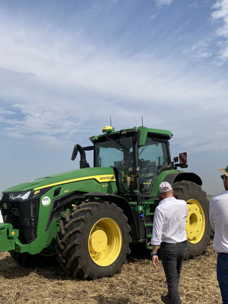 Két személy egy nagy John Deere traktort vizsgál meg egy mezőn, részben felhős égbolt alatt. A traktor zöld színű, sárga díszítéssel, és méretes gumiabroncsokkal rendelkezik, amelyeket terepmunkához terveztek. Az egyik ember fehér inget és sapkát visel, míg a másik öltözéke kevésbé látszik.