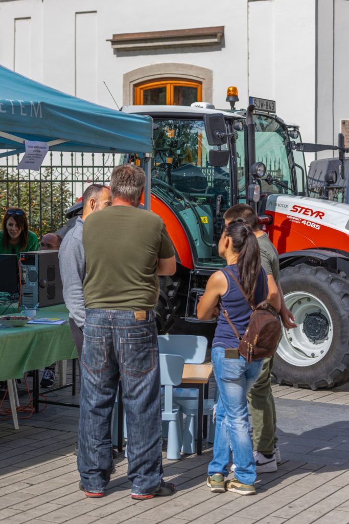 Az emberek egy sátor alatt gyülekeznek egy asztal körül elektronikus berendezésekkel. A közelben egy piros-fehér traktor parkol. A környezet a szabadban, egy ablakos és kerítésű épület közelében található. Úgy tűnik, hogy a csoport vitában vagy demonstrációban vesz részt.