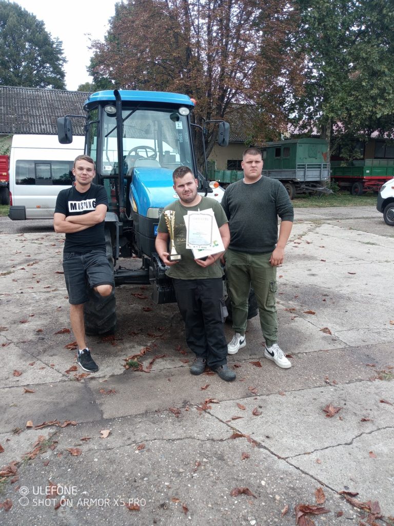 Három férfi áll egy kék traktor előtt. A középen álló férfi egy trófeát és egy oklevelet tart a kezében. A háttérben fák és parkoló járművek láthatók. A helyszín egy vidéki vagy farmszerű területnek tűnik.