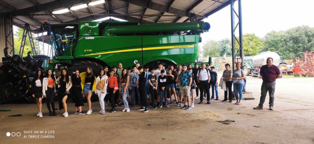 Emberek nagy csoportja pózol egy zöld John Deere 5770-es mezőgazdasági gép előtt, egy nagy nyitott fészerben. A csoport különböző korú, lezseren öltözött egyénekből áll a méretes gépezet előtt.