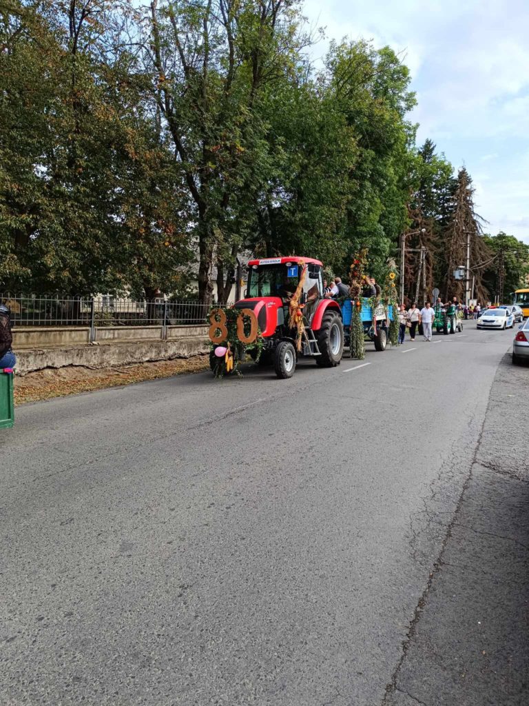 Zöldségből és virágokból készült nagy „80-as”-al díszített piros traktor vezet felvonulást. A traktort több úszó és ember követi egy fákkal szegélyezett utcán. A bámészkodók a járdákról nézik. Az esemény ünnepinek tűnik, alkalmat ünnepel.