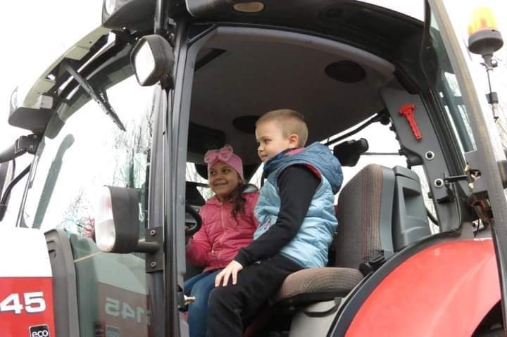 Két gyerek ül egy piros traktor vezetőfülkéjében. A rózsaszín kabátot és masnival ellátott fejpántot viselő lány mosolyogva várja. A kék mellényes, fekete inges fiú mellette ül, szintén mosolyogva. A traktor Massey Ferguson 5455 márkájú.