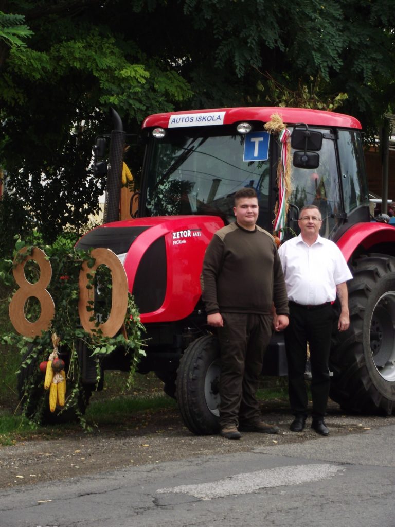 Két férfi áll egy piros traktor előtt, amelyet lombokkal és egy nagy "80" táblával díszítettek. A bal oldali férfi sötét munkaruhát, míg a jobb oldali férfi fehér inget és sötét nadrágot visel. A traktor szélvédőjén „AUTÓS ISKOLA.