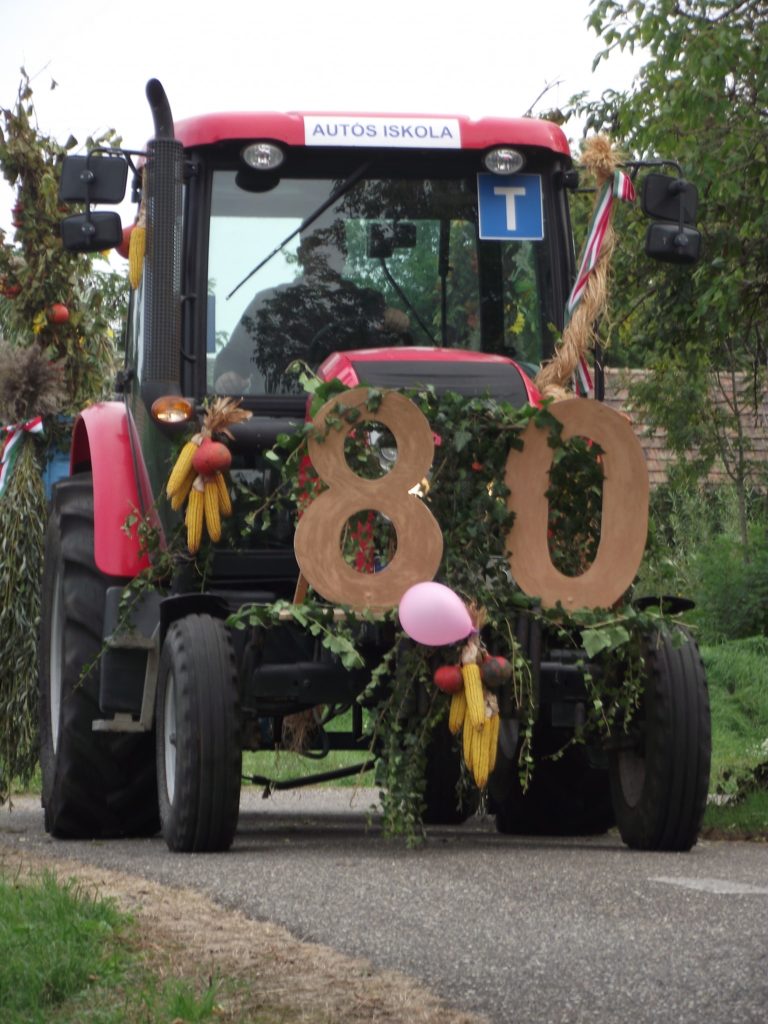 Egy vidéki úton áll egy piros traktor, amelyet lombozat és nagy, fából készült "80" számok díszítenek. A traktor szélvédőjén "AUTOS ISKOLA" felirat látható. Díszítő elemek, köztük kukorica és léggömbök vannak rögzítve a járműhöz. Fák és fű a háttérben.