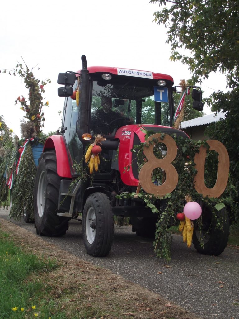 A nagy, 80-as, fából készült számokkal, kukoricahéjjal és növényzettel díszített piros traktort ünnepi díszítéssel vagy felvonulásra sugallják. A traktoron "AUTOS ISKOLA" felirat látható, és egy hasonlóan díszített pótkocsit húz maga mögött.