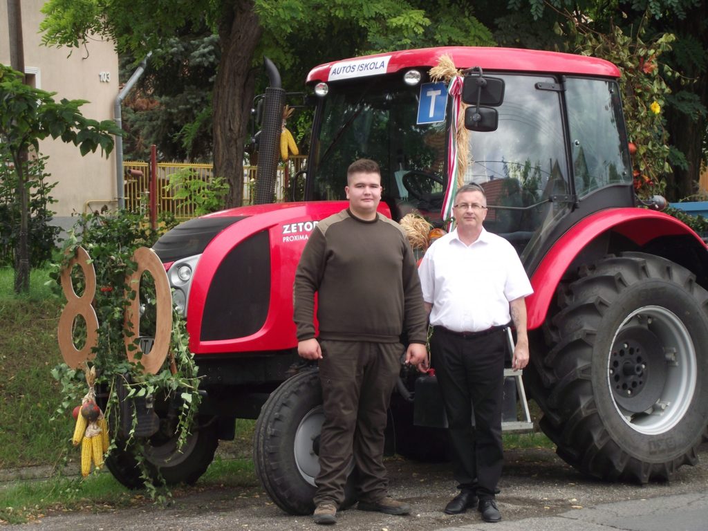 Két férfi áll egy díszített piros traktor előtt szabadtéri környezetben. Az egyik férfi fehér inget és fekete nadrágot, míg a másik barna ruhát visel. A traktort különféle díszítések díszítik, köztük nagy "80"-as kivágások. A háttérben fák és egy épület látható.
