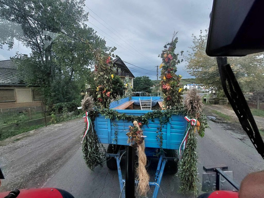 Egy kék pótkocsit vontat egy jármű egy vidéki úton. Az utánfutót zöld növények, virágok és szalagok díszítik. Az út két oldalán házak és fák állnak, az ég borús.