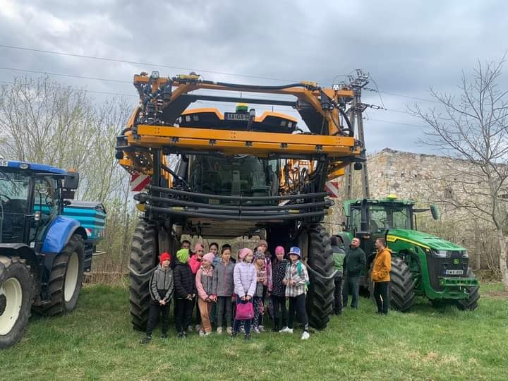 Egy csapat gyerek áll egy nagy mezőgazdasági gép előtt, mellette két traktor, egy kék és egy zöld. A szabadban egy füves mezőn vannak, fákkal és részben felhős égbolttal a háttérben.