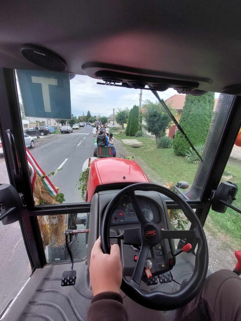 Kilátás egy traktor vezetőfülkéjéből, amelyen a vezető keze a kormányon és más járművek előtt látható egy külvárosi utcában. Az elöl haladó traktorhoz csatlakoztatott pótkocsi hátulján emberek láthatók.