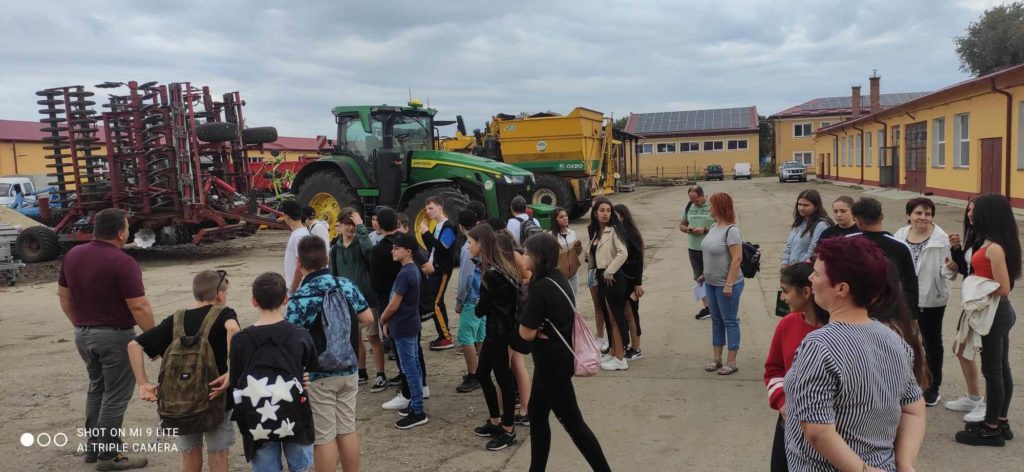 Emberek csoportja, köztük gyerekek és felnőttek, összegyűlik a szabadban, nagy mezőgazdasági gépek, köztük egy John Deere traktor és egyéb mezőgazdasági berendezések közelében. A háttérben a felhős ég alatt sárga és piros külsővel rendelkező épületek láthatók.