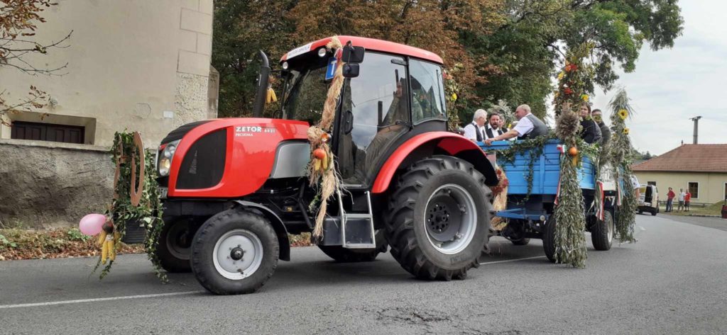 A kukoricaszárral és őszi levelekkel díszített piros Zetor traktor kék, zölddel és ünnepi díszekkel díszített pótkocsit húz. Az utánfutón bent ülők utaznak, akik részt vesznek egy helyi rendezvényen vagy egy falusi úton lévő felvonuláson.