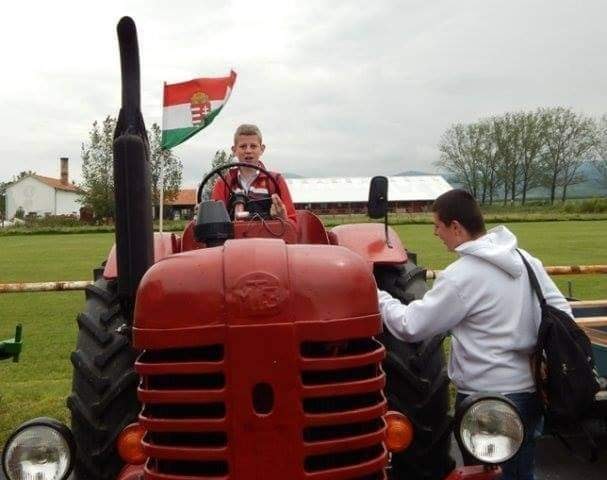 Egy fiatal fiú ül egy piros traktoron, magyar zászlóval, mellette egy fehér kabátot viselő férfi áll. Egy füves területen vannak, fákkal és épületekkel a háttérben a felhős ég alatt.