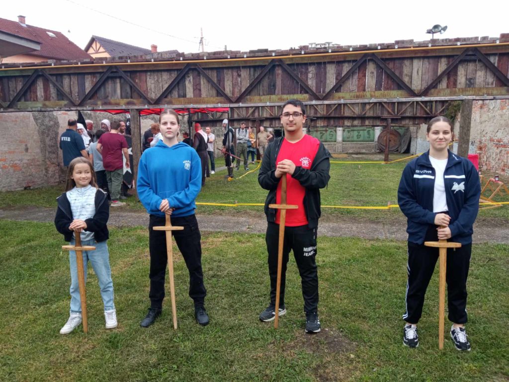 Négy fiatal, két fiú és két lány áll a füvön fakarddal. Mögöttük van egy szabadtéri terület faszerkezetekkel, történelmi jelmezű emberekkel és íjászcélokkal. Az időjárás borúsnak tűnik.