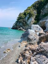 Festői partvonal tiszta kékeszöld vízzel, sziklás stranddal és egy tengerbe nyúló, erdős sziklával. Az ég többnyire tiszta, néhány szeles felhővel, kiemelve a tengerparti táj nyugodt és természetes szépségét.